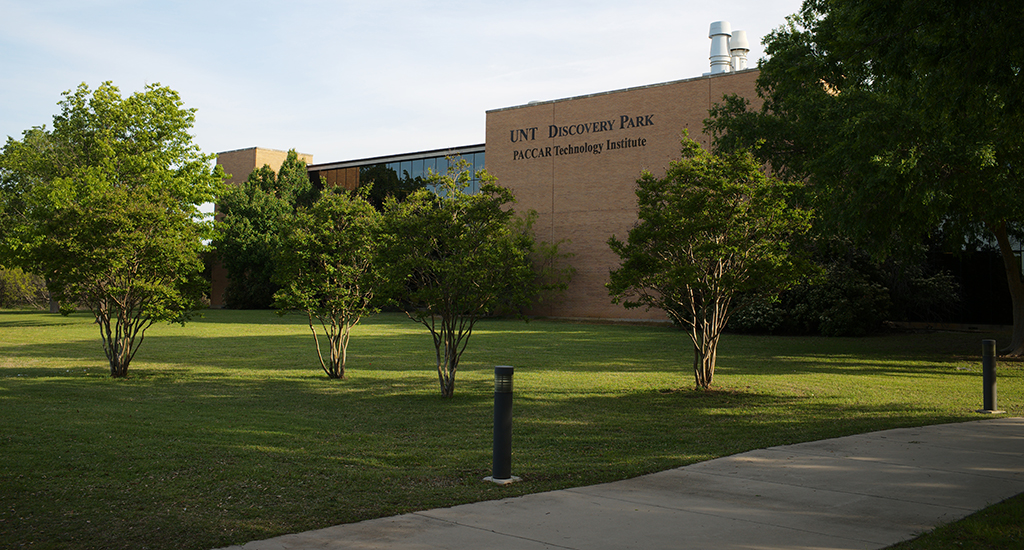 Photo features a building surrounded with trees that includes the words "UNT Discovery Park" and "PACCAR Technology Institute" on wall