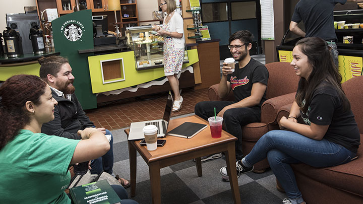 Four students sit around a coffee table chatting with drinks in hands
