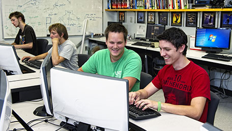 Two students look at computer screen smiling with another two at background.