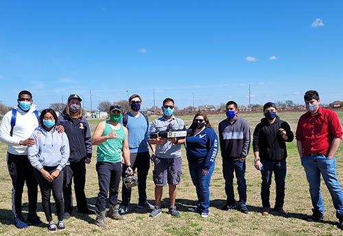 Ten students stand in a line outside with masks and pose while holding a drone and drone controller