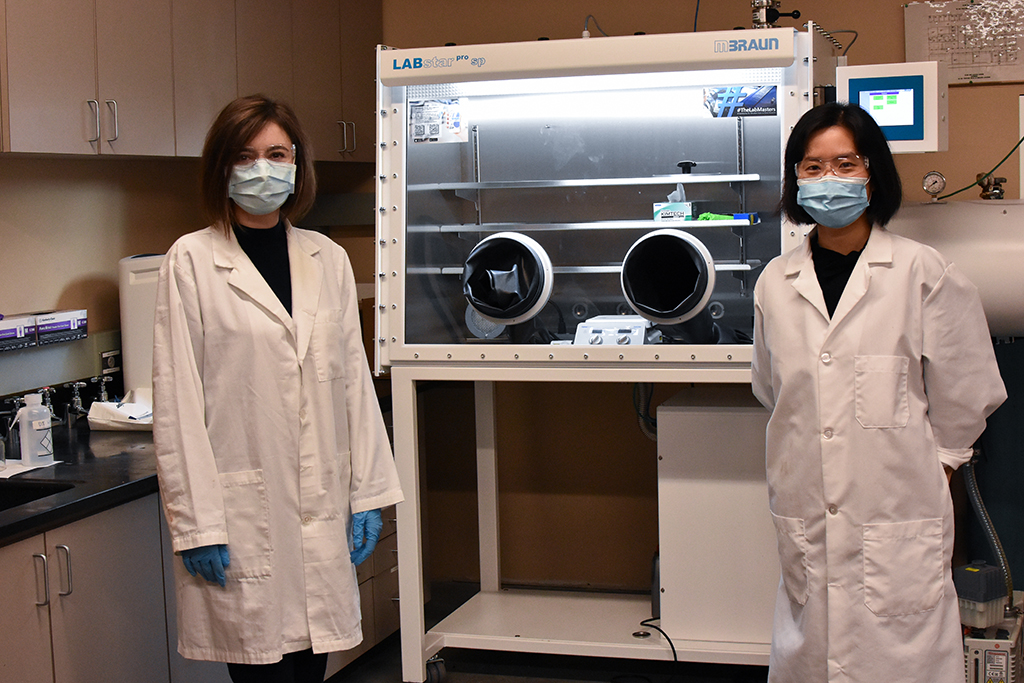 Xiao Li and a graduate student stand in front of machinery with lab coats and safety goggles