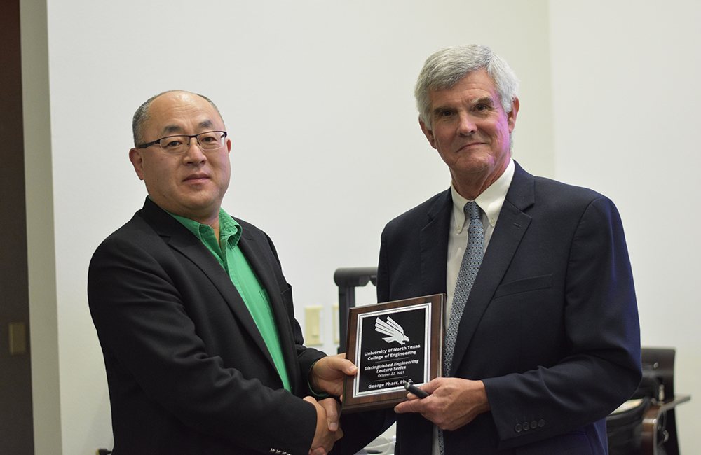 Dean Hanchen Huang stands on the left shaking hands with George Pharr on the right. A plaque is in between them that honors George Pharr.